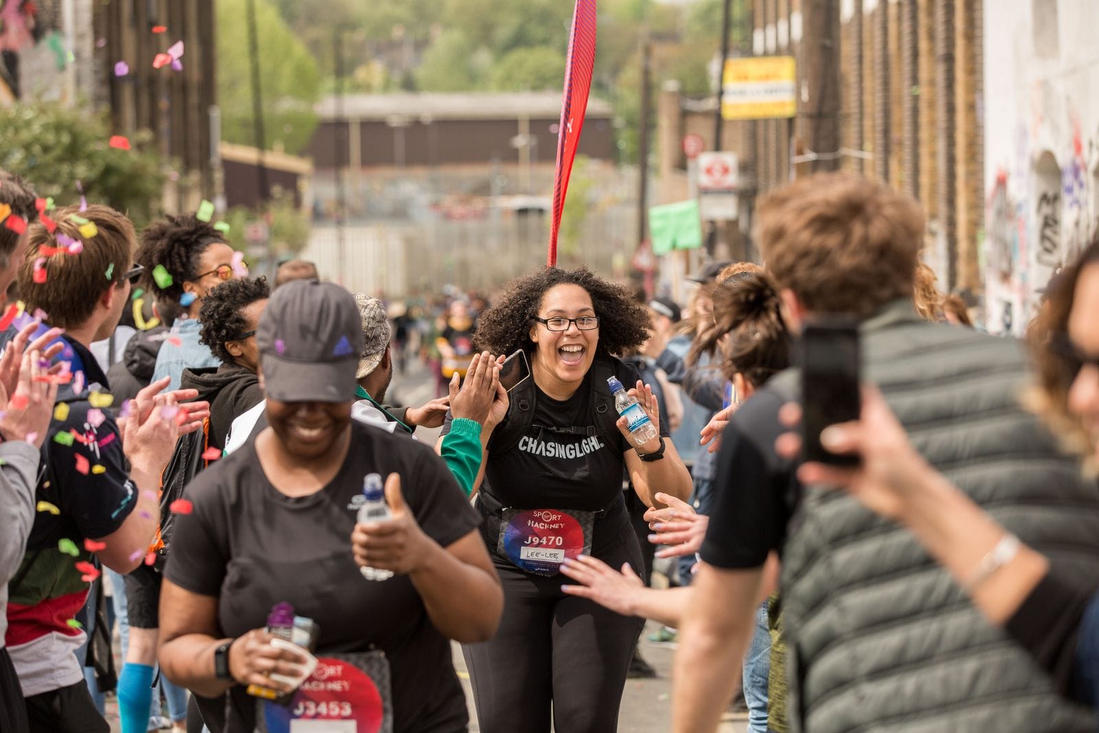 A Black British woman crosses the finish line at the Hackney Half Marathon in London in a shower of confetti while spectators cheer her on.
